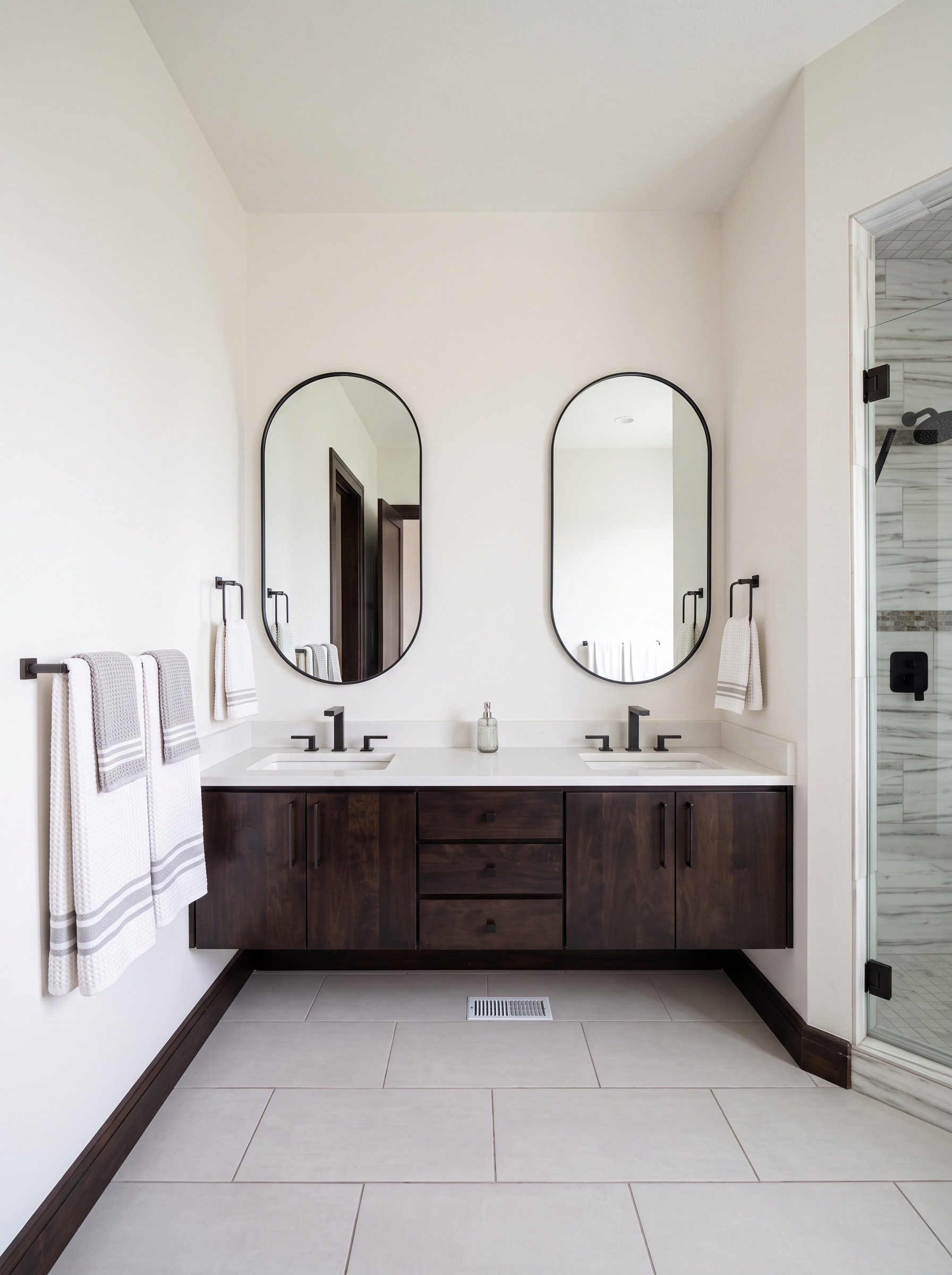 Double vanity with arched mirrors, espresso cabinets, and marble-look tile shower